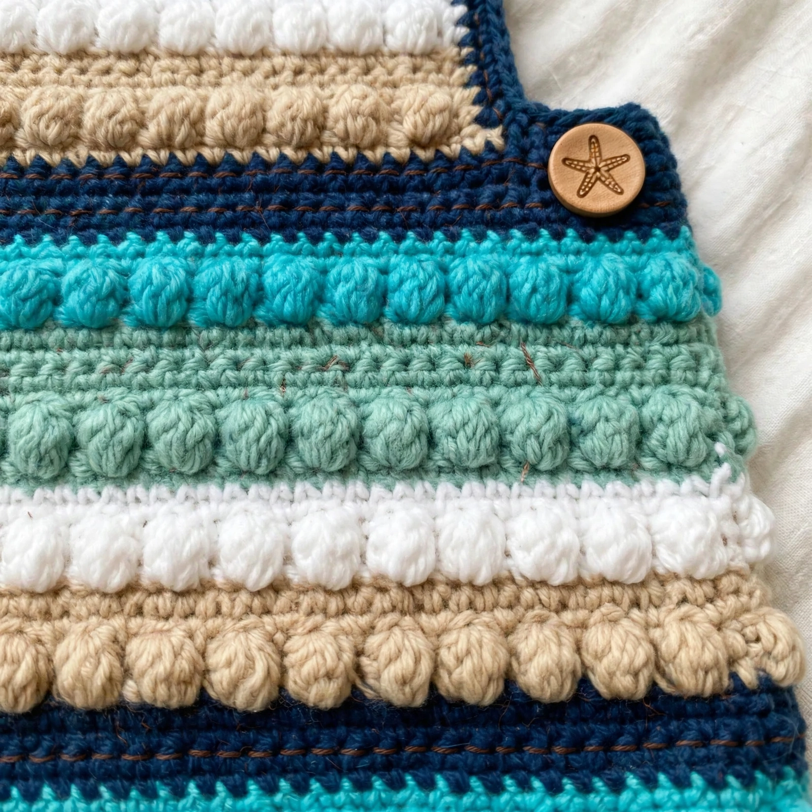 Close-up of the bobble-textured crochet stripes in teal, sage, white and tan with a wooden starfish button